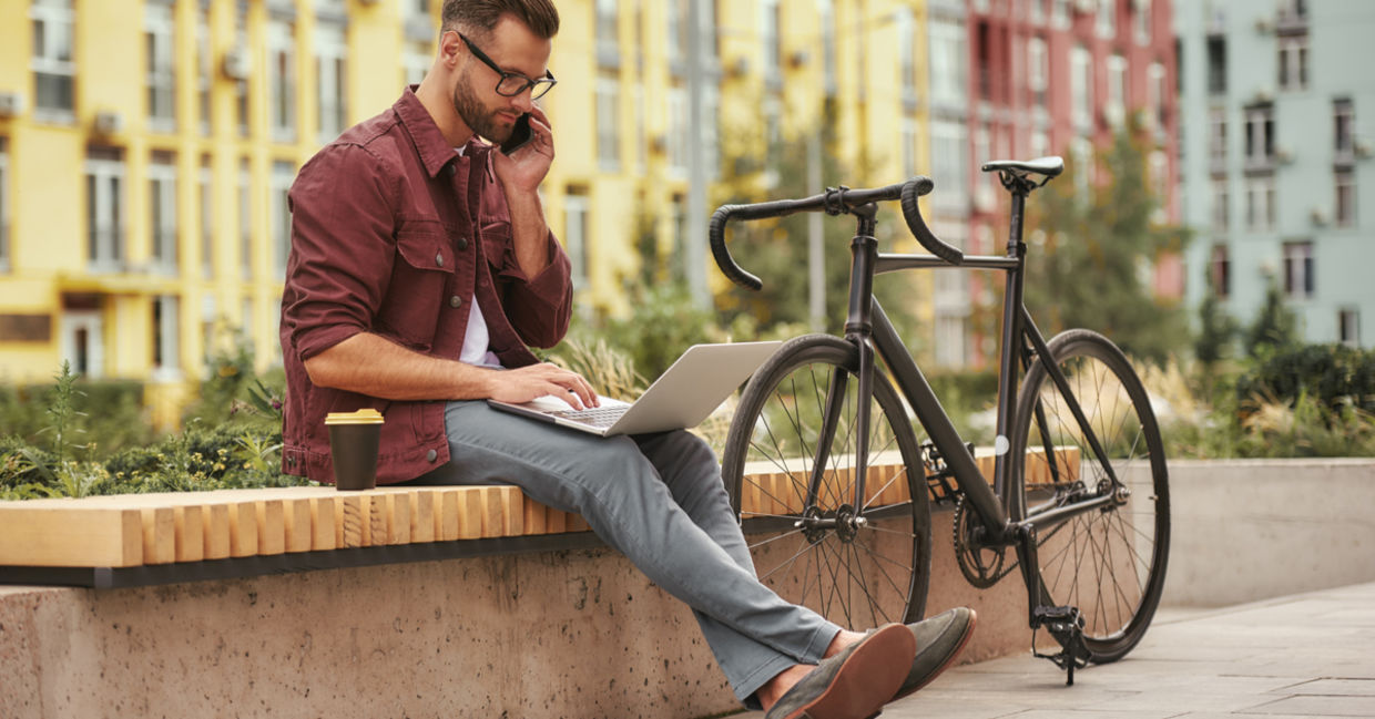 Young man working on laptop and talking by phone while sitting on the bench near his bicycle