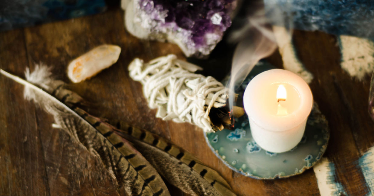A sage smudge stick, amethyst, a feather, and a lit candle create a sacred space for meditation.