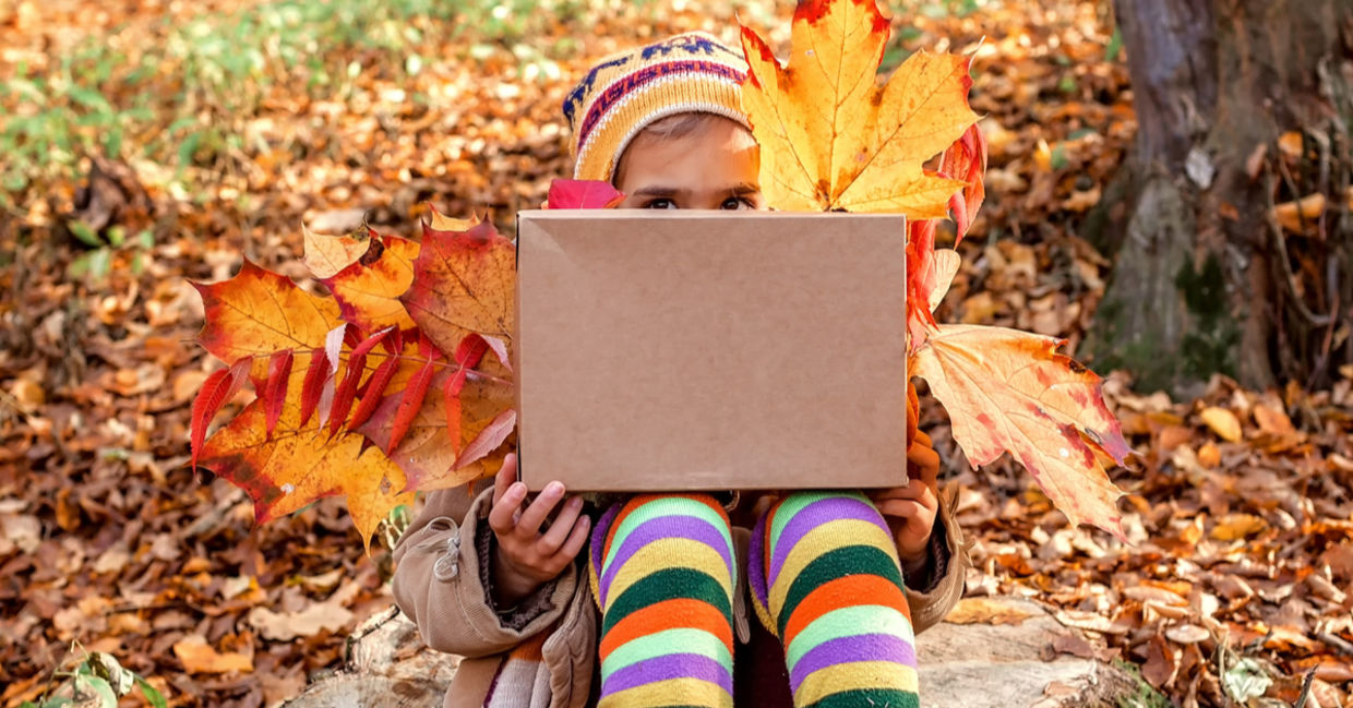 A young girl holding autumn leaves peeks from behind a large gift box.