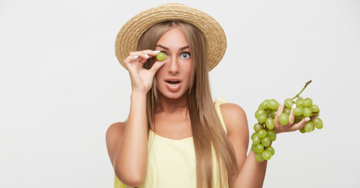 A young woman holds a green grape in front of her eye.