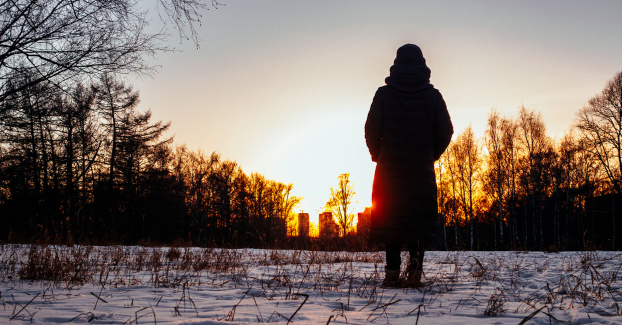 A woman stands among trees in the snow watching the sun setting behind buildings.