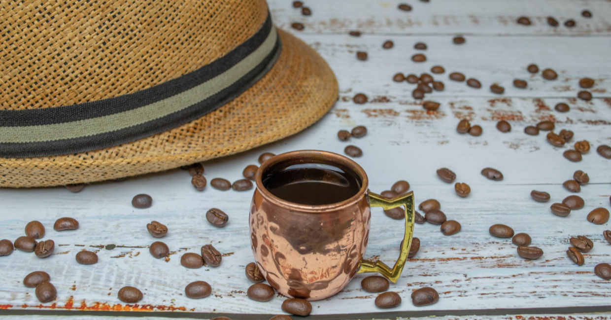 Copper cup with espresso on white wooden table with organic coffee beans with Cuban hat in the background.