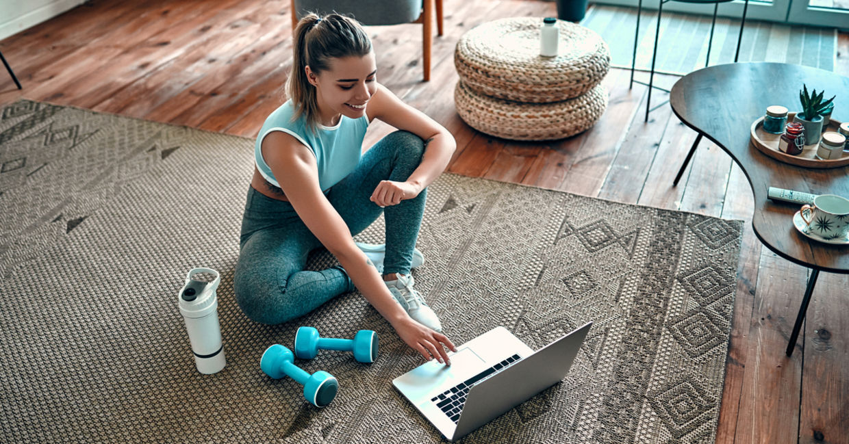 Sporty woman sitting on the floor with sports equipment around her as she uses a laptop.