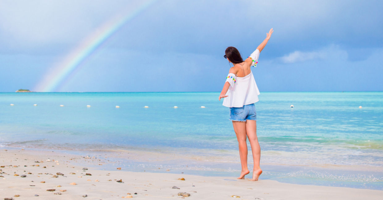 A young woman joyfully walks along a beach with a rainbow on the horizon.