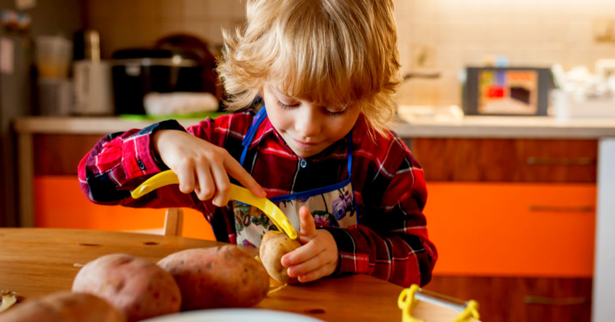 A little kid is carefully peeling potatoes in her kitchen.