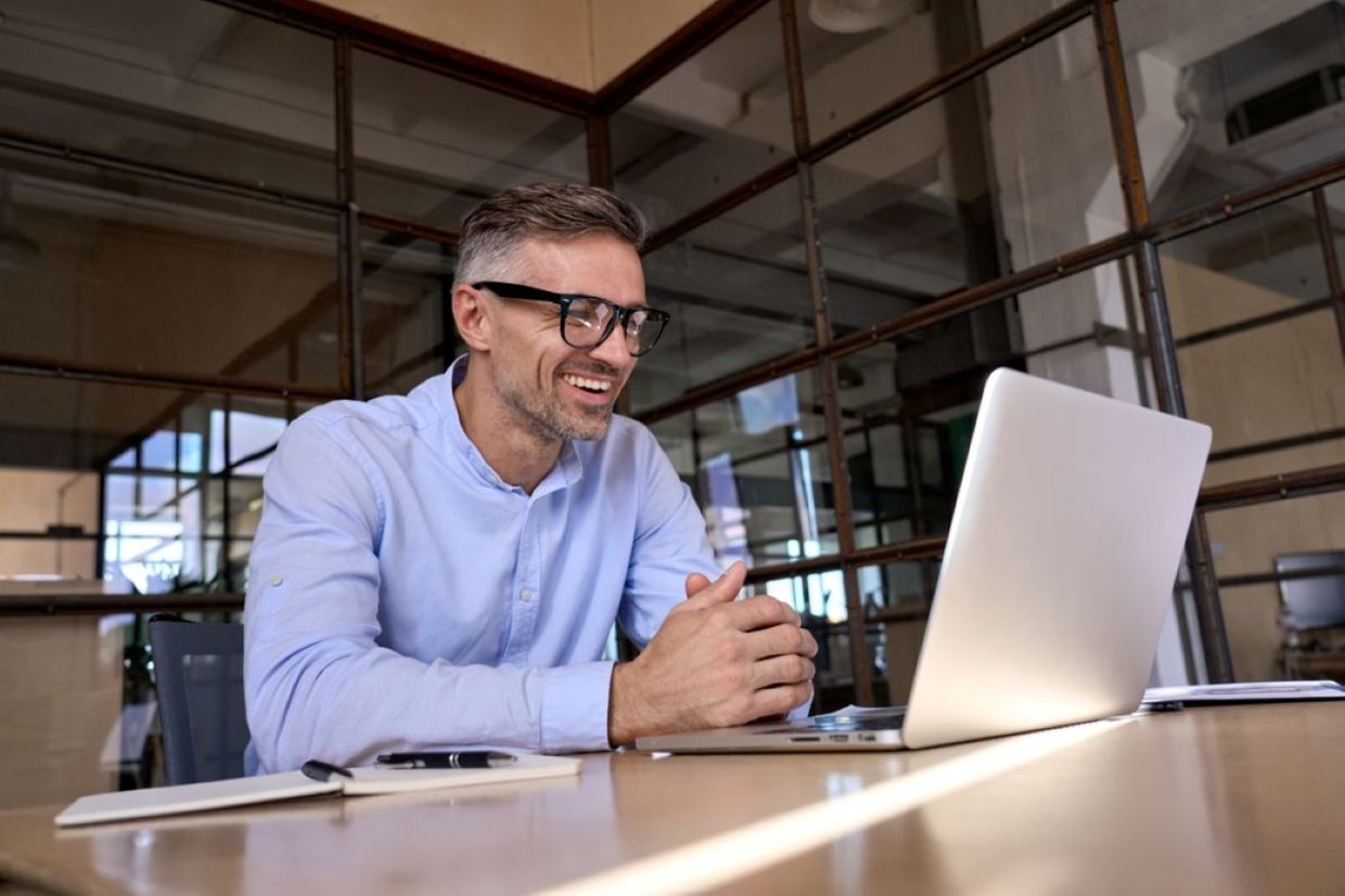 A man wearing glasses smiles while looking at a laptop screen.