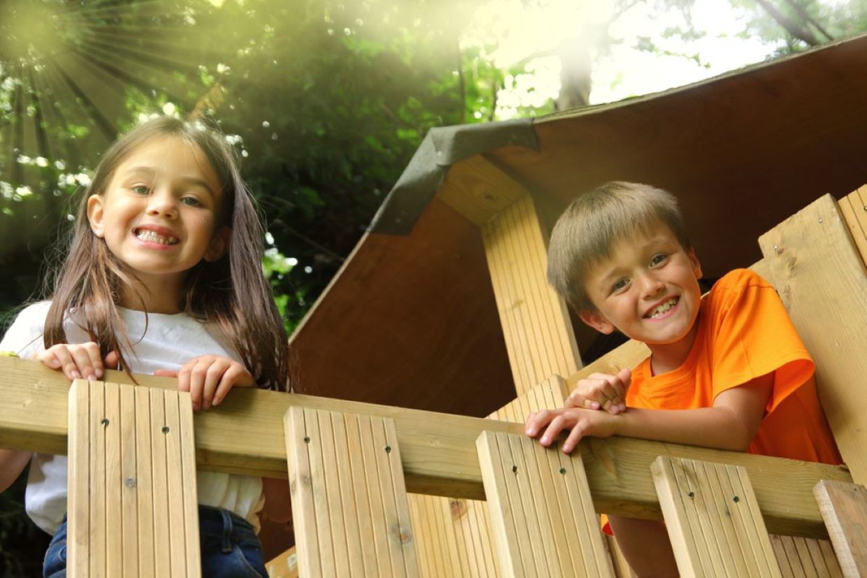 Smiling children look out from a wooden treehouse.