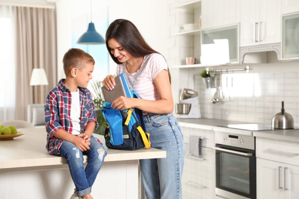 A smiling mother helps her son pack a backpack.