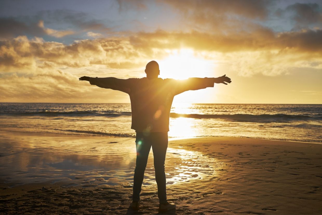 A silhouette of a man at the beach standing with his arms raised.