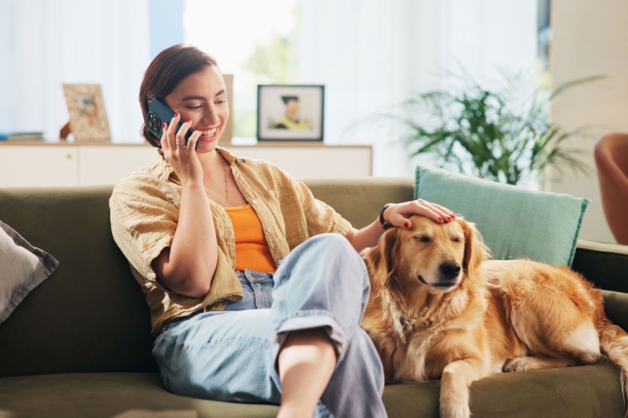 A woman sits on a sofa petting a dog while talking on the phone.