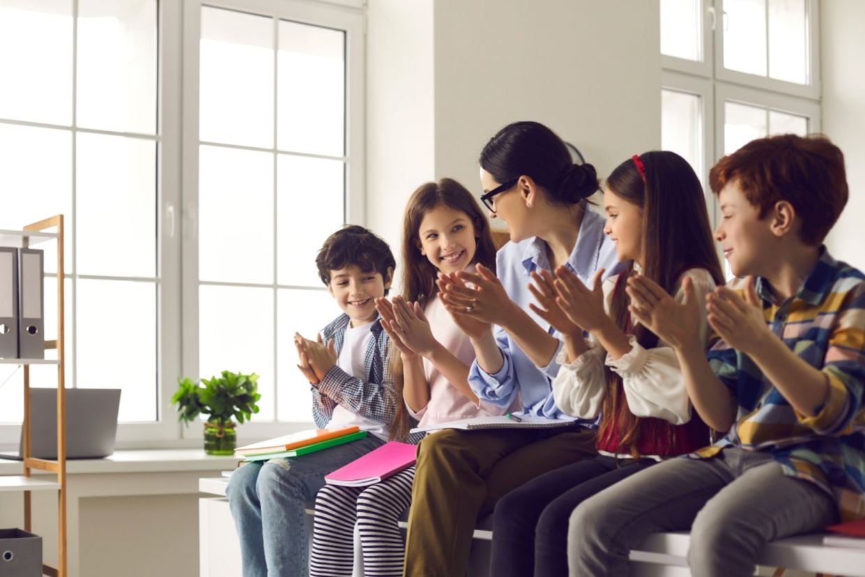 A female teacher claps together with a group of happy children.