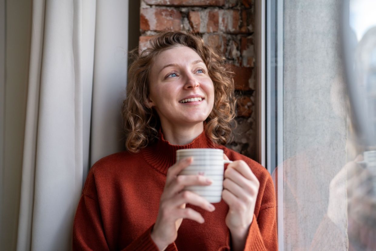 A smiling woman next to a window holds a mug.