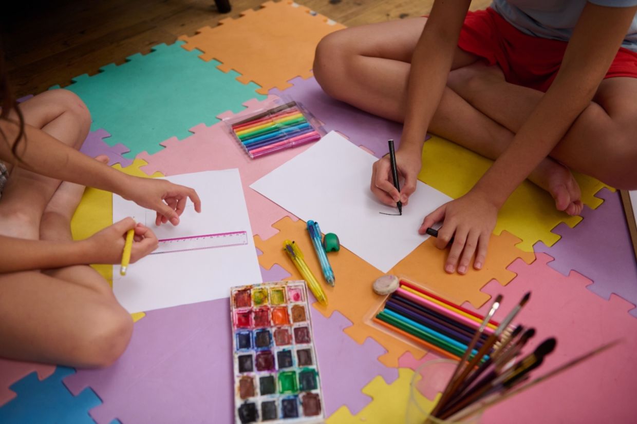 Two children sit on colorful mats and use art supplies.
