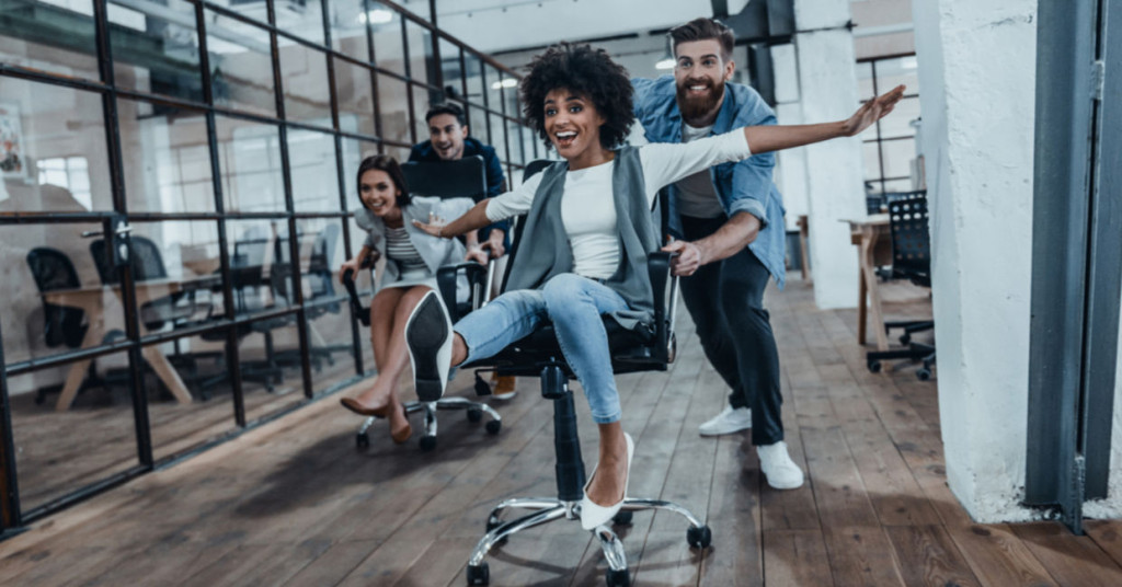Group of coworkers racing on office chairs in a modern open-plan office, with one person in the foreground celebrating with arms outstretched.