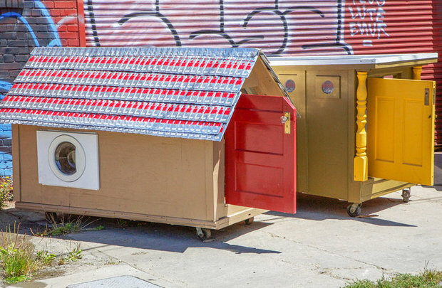 Row of small cardboard box houses with colorful roofs in red and blue checkered patterns, featuring red and yellow doors, with a washing machine visible on one unit.