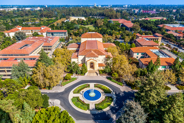 Stanford Aerial view of stanford university campus with a domed central building, circular plaza with blue fountain, surrounded by green trees and orange-roofed buildings.