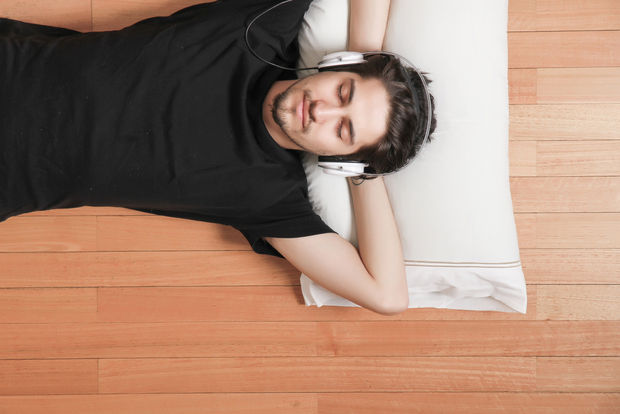 Music Person lying on a wooden floor wearing headphones around their neck, relaxing and smiling while resting on a white pillow.