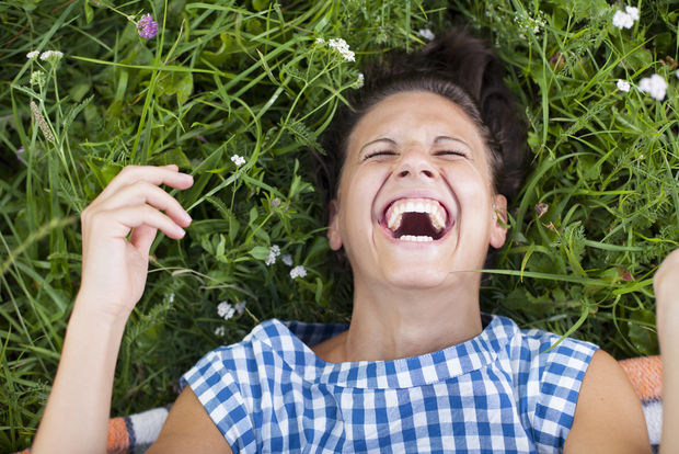 Laughter Person laughing while lying on their back in grass and wildflowers, wearing a blue and white checkered shirt.