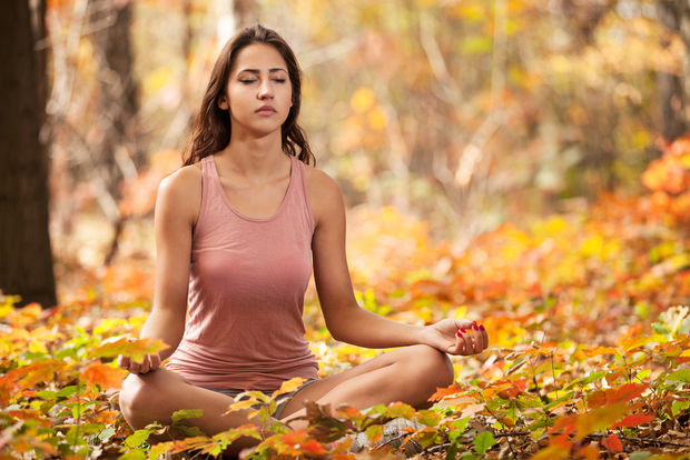 Woman in pink tank top sitting in meditation pose surrounded by orange and yellow fall leaves in a forest.