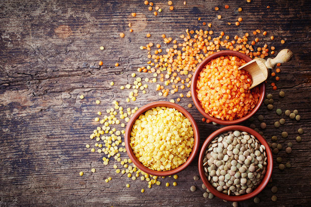 photo Three terracotta bowls containing different varieties of lentils — red lentils, yellow split lentils, and green/brown lentils — arranged on a dark rustic wooden surface with a wooden scoop and scattered lentils.
