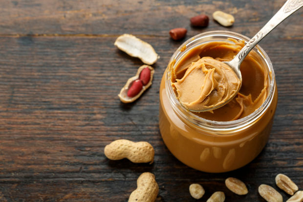 photo An open glass jar of creamy peanut butter with a silver spoon scooping out a dollop, surrounded by whole and shelled peanuts on a dark wooden surface