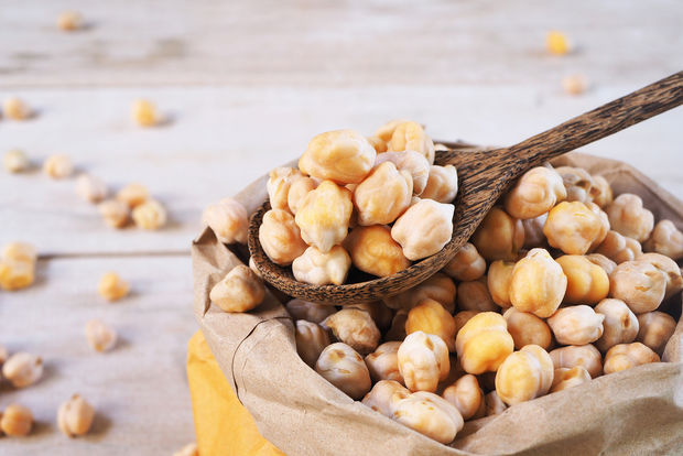 photo Dried chickpeas spilling from a brown paper bag with a wooden spoon scooping them up, on a light wooden surface with scattered chickpeas in the background.