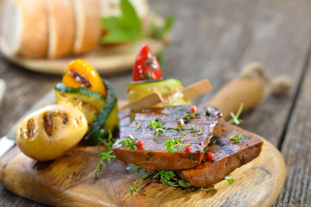 photo Grilled marinated seitan steaks garnished with fresh thyme and pink peppercorns, served on a round wooden board with a colorful vegetable skewer and grilled onions, with sliced bread in the background.