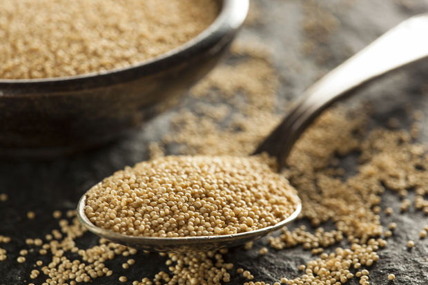 photo A silver spoon overflowing with tiny golden amaranth seeds in the foreground, with a rustic metal bowl filled with more amaranth in the background, on a dark slate surface with scattered seeds.