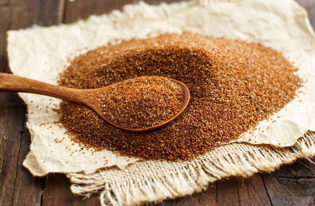 photo A pile of reddish-brown teff grain spread on a cream-colored linen cloth, with a wooden spoon resting in the center, on a dark rustic wooden table.
