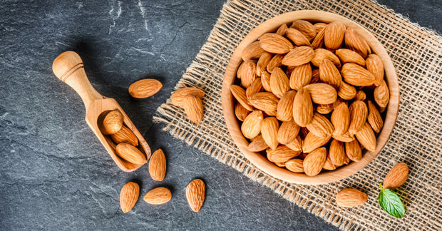 photo A wooden bowl filled with whole almonds and a small wooden scoop on burlap cloth over a dark slate surface.
