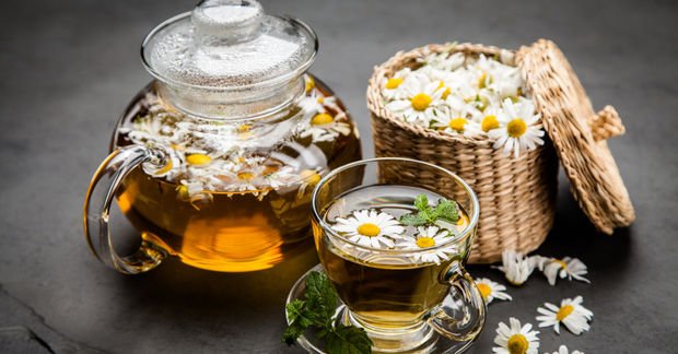 photo A glass teapot and cup of chamomile tea with floating daisies, beside a woven basket of fresh chamomile flowers.