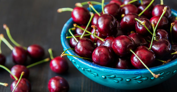 photo Fresh dark red cherries with green stems overflowing from a turquoise ceramic bowl on a dark wooden surface.