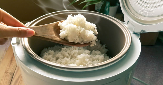 photo A hand lifts a wooden spoon of steaming white rice from an open rice cooker, with steam rising from the pot.