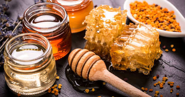 photo Honeycomb, jars of different honey varieties, a wooden dipper, and bee pollen on a dark slate surface.