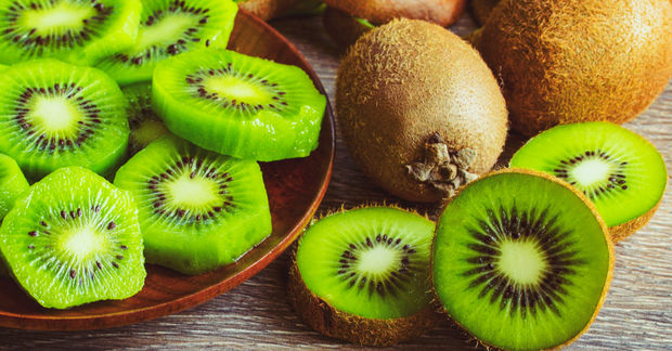 photo Sliced green kiwi fruit on a wooden plate alongside whole and halved kiwis on a light wooden surface.
