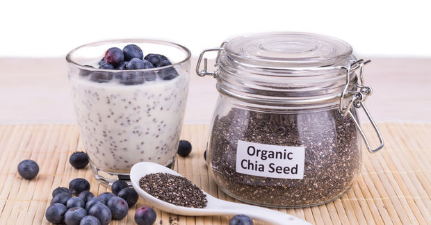 photo A glass jar labeled 'Organic Chia Seed' with a metal clasp lid, alongside a glass of chia seed pudding topped with fresh blueberries, a white ceramic spoon filled with chia seeds, and scattered blueberries on a bamboo mat.