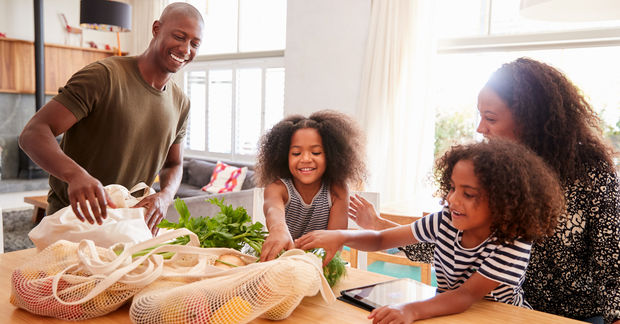photo A happy family unpacks fresh groceries from reusable bags onto a kitchen table in a bright living space.