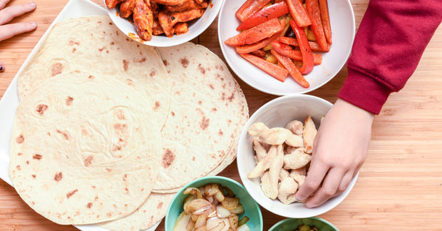 photo Child's hand reaching for cashews while assembling a wrap or taco with tortilla, cooked bacon, red bell peppers, and other ingredients on a wooden surface.