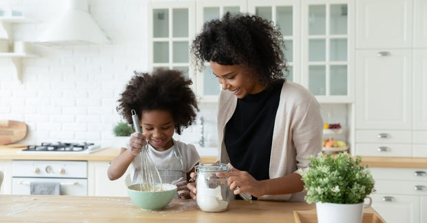 photo Woman and child baking together in a bright white kitchen, with the child wearing an apron and stirring a bowl of ingredients.