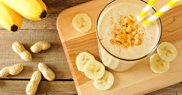 It Banana peanut butter smoothie topped with granola in a glass, surrounded by banana slices, whole peanuts, and fresh bananas on a wooden board.