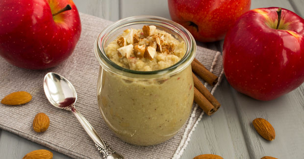This Glass jar of apple cinnamon yogurt topped with almond slices, surrounded by fresh red apples, almonds, cinnamon sticks, and a spoon on a white cloth.