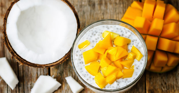 Who Bowl of coconut chia seed pudding topped with fresh mango chunks, with a coconut half and fresh mango visible alongside on a wooden surface.