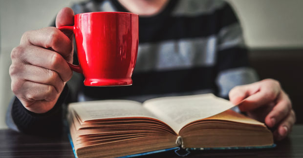 Person holding a bright red mug while reading an open book, with the mug in sharp focus and the reader blurred in the background.