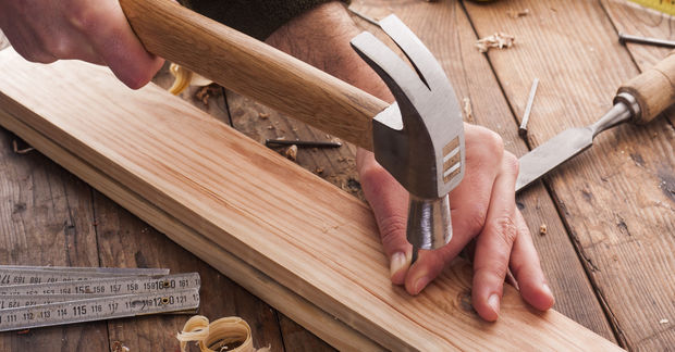 Hands using a hammer to drive a nail into a wooden board surrounded by woodworking tools and measuring tape on a wooden work surface.