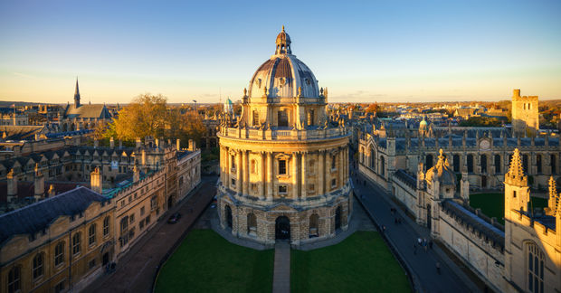 University An aerial view of the University of Oxford, Aerial view of a grand domed classical stone building illuminated by golden hour sunlight, surrounded by historic architecture and green courtyard, with church spires visible in background.