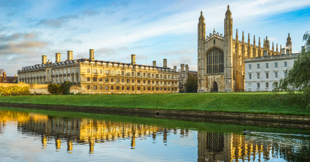 University Cambridge in all its glory, Historic university college buildings with Gothic chapel spire reflected in a river, featuring golden stone architecture and green lawns.