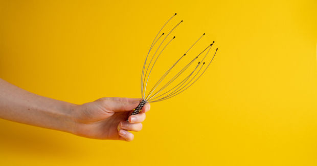 A A hand holds a wire scalp massager with metal prongs tipped in small balls against a bright yellow background.