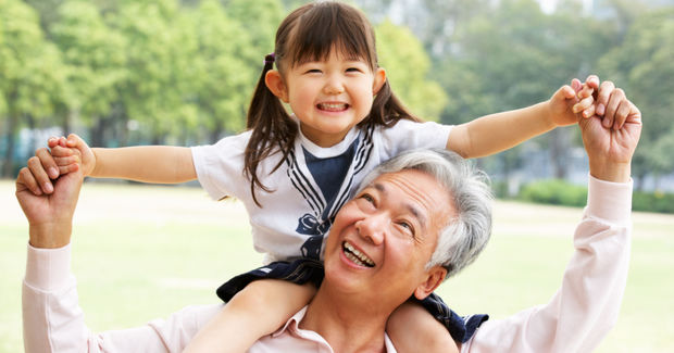 A Grandfather and granddaughter laughing together outdoors, with child sitting on his shoulders with arms spread wide in a park setting.