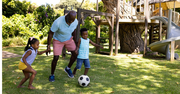 Teaching Man playing soccer with two young children in a sunny backyard with grass and wooden play structures.