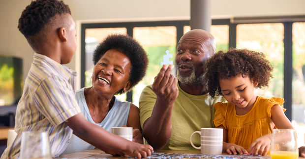 Doing Multi-generational family laughing together around a table playing a board game with mugs and natural light from windows in the background.